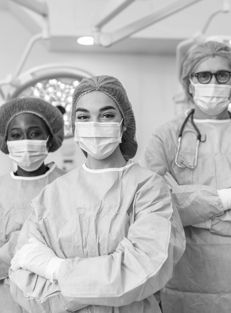 three clinicians in an operating room wearing sterile surgical gowns, gloves, and hair caps