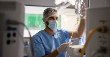 An anesthesiologist in blue scrubs and a surgical mask adjusts an IV bag in a hospital operating room equipped with medical machinery.