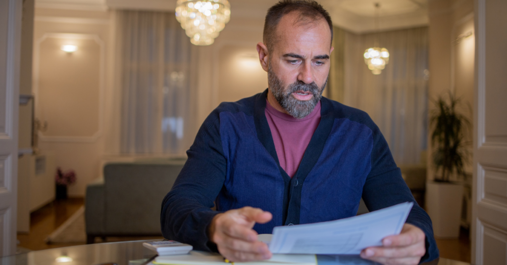 Mature male wearing a blue cardigan sweater sitting at a table inside a home reviewing tax documents