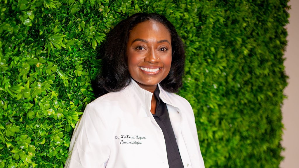 Anesthesiologist Dr. LaKesha Legree smile and wearing a white lab coat with a wall of plants behind her 