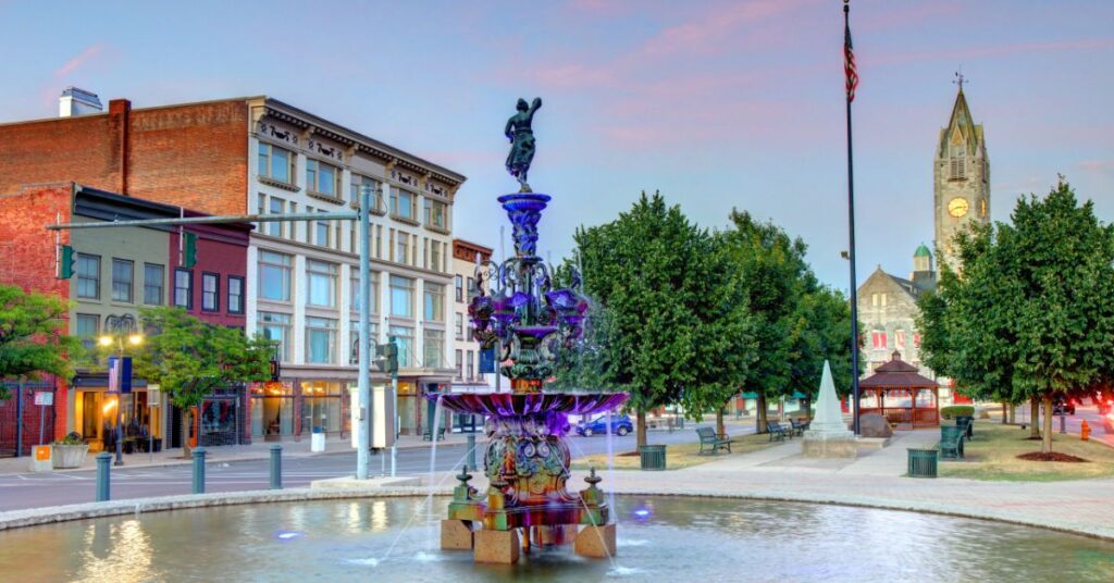 view of downtown Watertown, New York with a water fountain in the foreground. A flagpole with the U.S. flag is behind it and further back is a tall church building