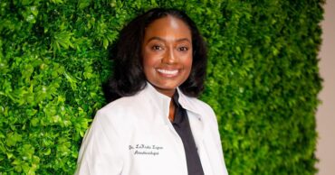 LaKesha Legree, an anesthesiologist dedicated to rural medicine poses in white coat in front of a green plant wall