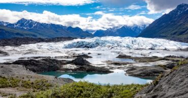 A glacier in Alaska stretches across a valley with a lake and rocky terrain in the foreground, surrounded by snow-capped mountains under a partly cloudy sky.